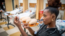 A nursing student draws medicine from a bottle into a syringe in front of the nursing classroom facilities with practice mannequins in hospital beds.