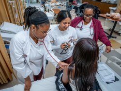 Two nursing students practicing with stethoscopes on a patient as the professor supervises.