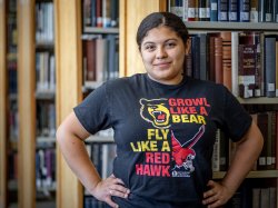 Odyeli Ramos Tobar stands in the Bloomfield library wearing a T-shirt with the Bloomfield and Montclair mascots, a bear and red hawk.